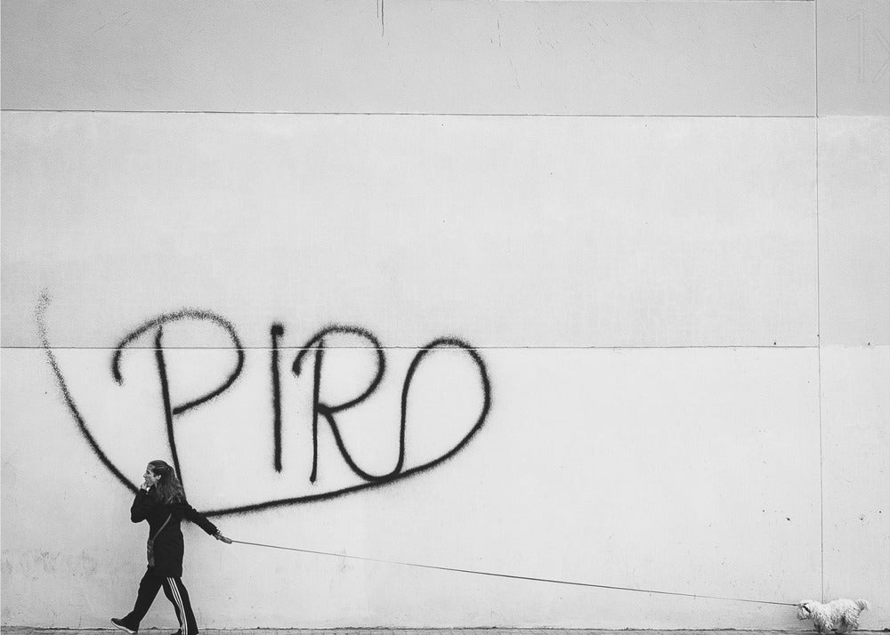 A minimalist black and white photograph of a woman dressed in black walking a small, fluffy white dog on a long leash. They are walking along a sidewalk in front of a large, pale wall with the word 'PIRO' written in black spray paint graffiti. The entire image is enclosed in a light grey frame. Poster