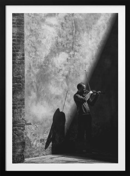 A dramatic black and white photograph of a man playing the violin, standing in a corner against a weathered wall. He is bathed in a sharp diagonal ray of light, casting a long, dark shadow behind him. Artwork