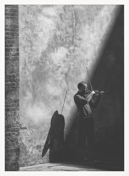 A dramatic black and white photograph of a man playing the violin, standing in a corner against a weathered wall. He is bathed in a sharp diagonal ray of light, casting a long, dark shadow behind him. Artwork