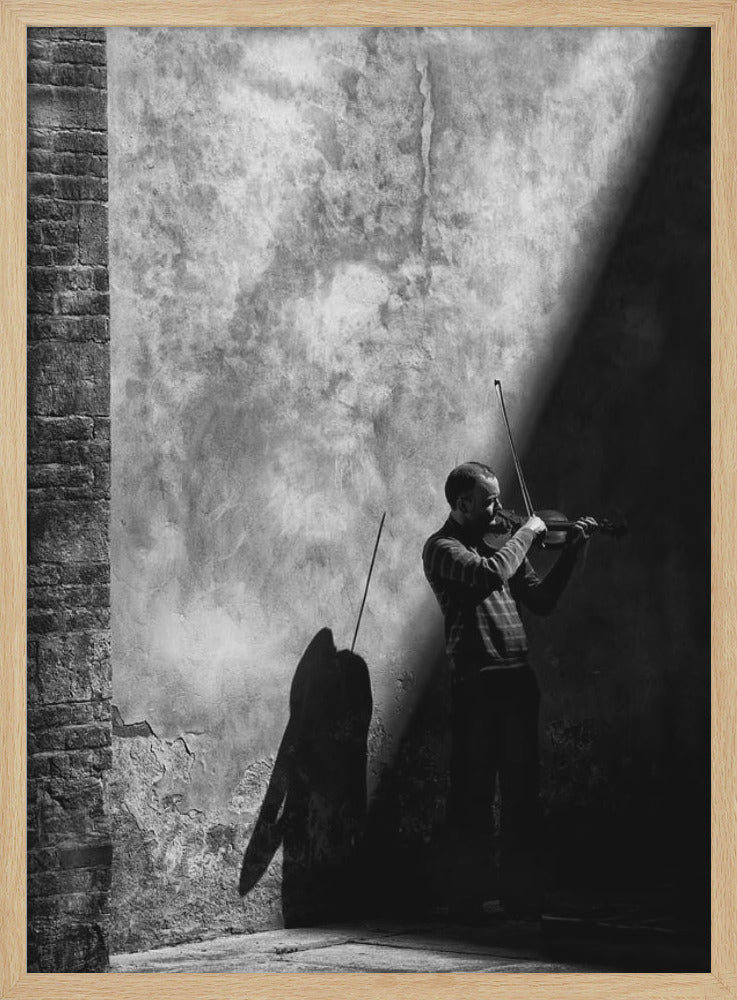 A dramatic black and white photograph of a man playing the violin, standing in a corner against a weathered wall. He is bathed in a sharp diagonal ray of light, casting a long, dark shadow behind him. Artwork