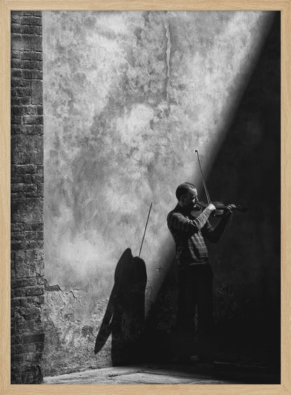 A dramatic black and white photograph of a man playing the violin, standing in a corner against a weathered wall. He is bathed in a sharp diagonal ray of light, casting a long, dark shadow behind him. Artwork