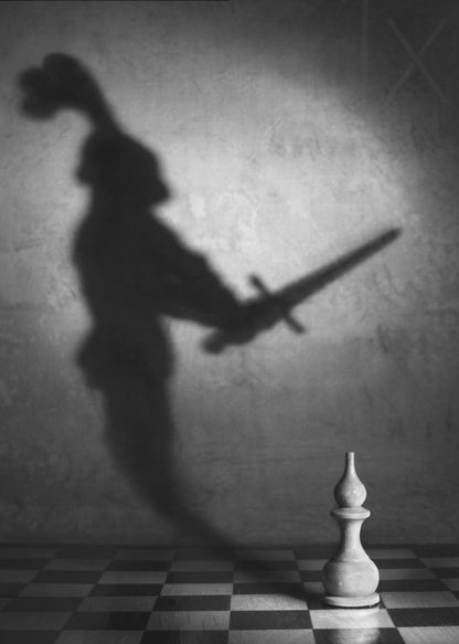 A black and white conceptual photograph of a single white chess piece on a checkerboard, casting a large, ominous shadow of a medieval knight holding a sword against a textured wall. Print