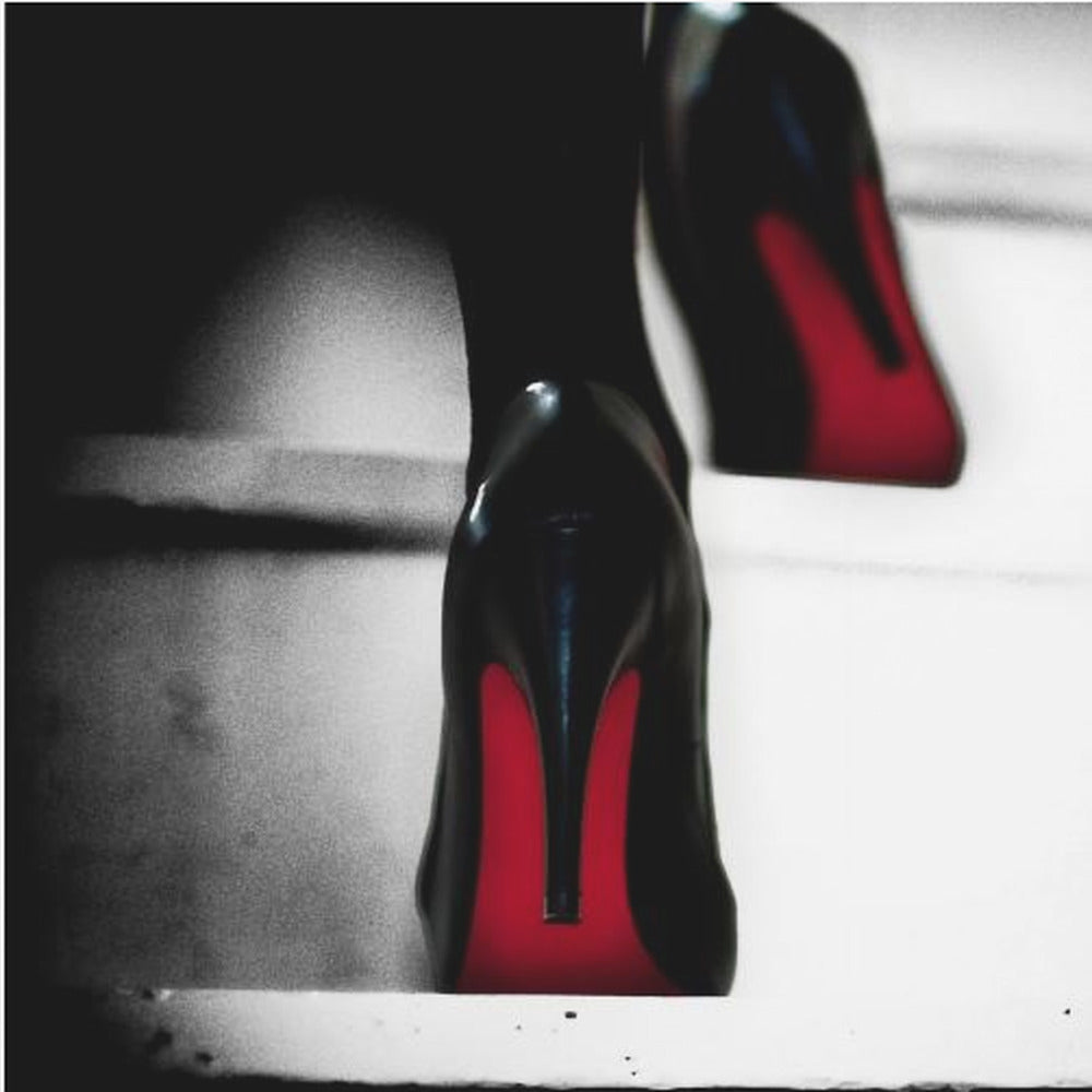A dramatic, low-angle, black and white photograph focusing on the back of a person wearing glossy black high heels with vibrant red soles, all set within a light wood frame. Artwork