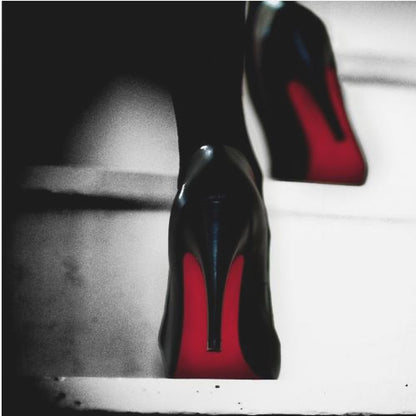 A dramatic, low-angle, black and white photograph focusing on the back of a person wearing glossy black high heels with vibrant red soles, all set within a light wood frame. Artwork