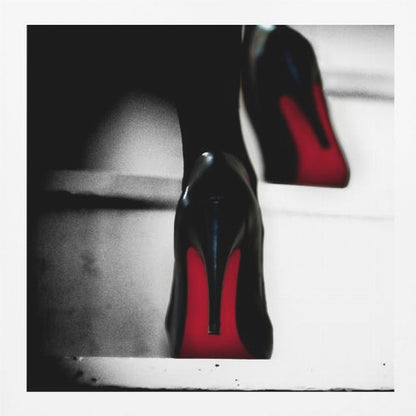 A dramatic, low-angle, black and white photograph focusing on the back of a person wearing glossy black high heels with vibrant red soles, all set within a light wood frame. Artwork