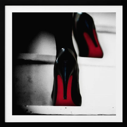 A dramatic, low-angle, black and white photograph focusing on the back of a person wearing glossy black high heels with vibrant red soles, all set within a light wood frame. Artwork
