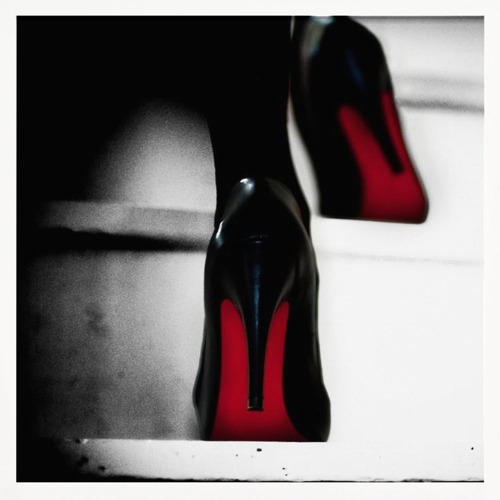 A dramatic, low-angle, black and white photograph focusing on the back of a person wearing glossy black high heels with vibrant red soles, all set within a light wood frame. Artwork