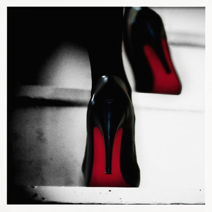 A dramatic, low-angle, black and white photograph focusing on the back of a person wearing glossy black high heels with vibrant red soles, all set within a light wood frame. Artwork