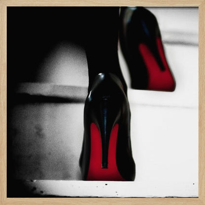 A dramatic, low-angle, black and white photograph focusing on the back of a person wearing glossy black high heels with vibrant red soles, all set within a light wood frame. Artwork