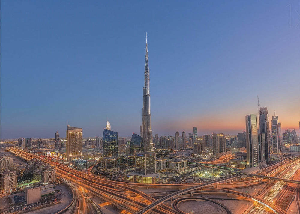 An aerial photograph of the Dubai skyline at dusk, centered on the illuminated Burj Khalifa. The sky transitions from deep blue to warm orange at the horizon, while a complex network of highways below glows with the light trails of traffic. Artwork