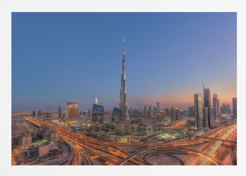 An aerial photograph of the Dubai skyline at dusk, centered on the illuminated Burj Khalifa. The sky transitions from deep blue to warm orange at the horizon, while a complex network of highways below glows with the light trails of traffic. Artwork
