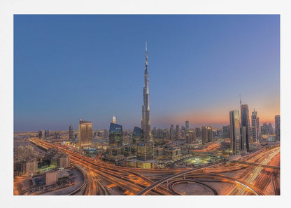 An aerial photograph of the Dubai skyline at dusk, centered on the illuminated Burj Khalifa. The sky transitions from deep blue to warm orange at the horizon, while a complex network of highways below glows with the light trails of traffic. Artwork