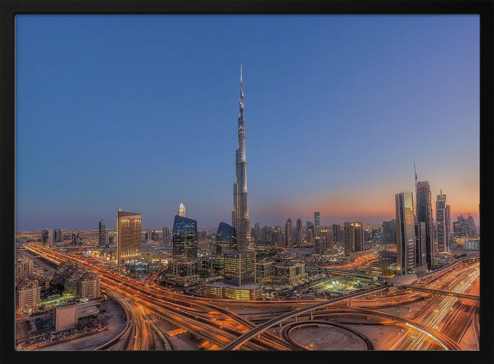 An aerial photograph of the Dubai skyline at dusk, centered on the illuminated Burj Khalifa. The sky transitions from deep blue to warm orange at the horizon, while a complex network of highways below glows with the light trails of traffic. Artwork