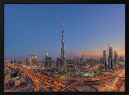 An aerial photograph of the Dubai skyline at dusk, centered on the illuminated Burj Khalifa. The sky transitions from deep blue to warm orange at the horizon, while a complex network of highways below glows with the light trails of traffic. Artwork