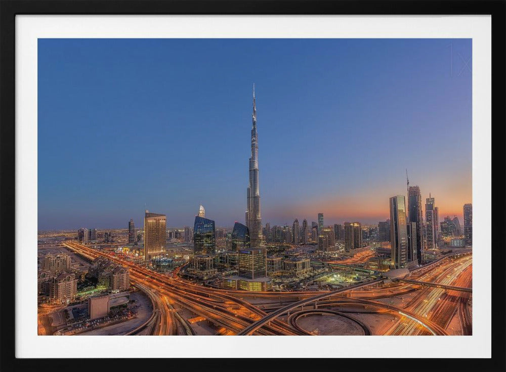 An aerial photograph of the Dubai skyline at dusk, centered on the illuminated Burj Khalifa. The sky transitions from deep blue to warm orange at the horizon, while a complex network of highways below glows with the light trails of traffic. Artwork