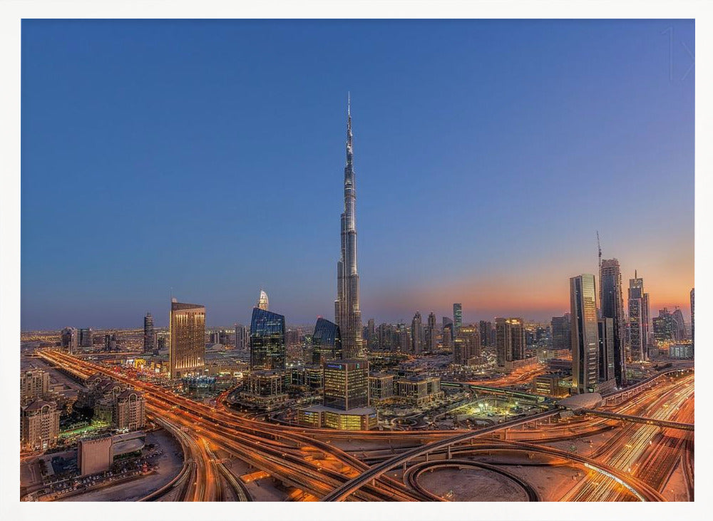 An aerial photograph of the Dubai skyline at dusk, centered on the illuminated Burj Khalifa. The sky transitions from deep blue to warm orange at the horizon, while a complex network of highways below glows with the light trails of traffic. Artwork