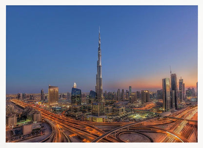 An aerial photograph of the Dubai skyline at dusk, centered on the illuminated Burj Khalifa. The sky transitions from deep blue to warm orange at the horizon, while a complex network of highways below glows with the light trails of traffic. Artwork