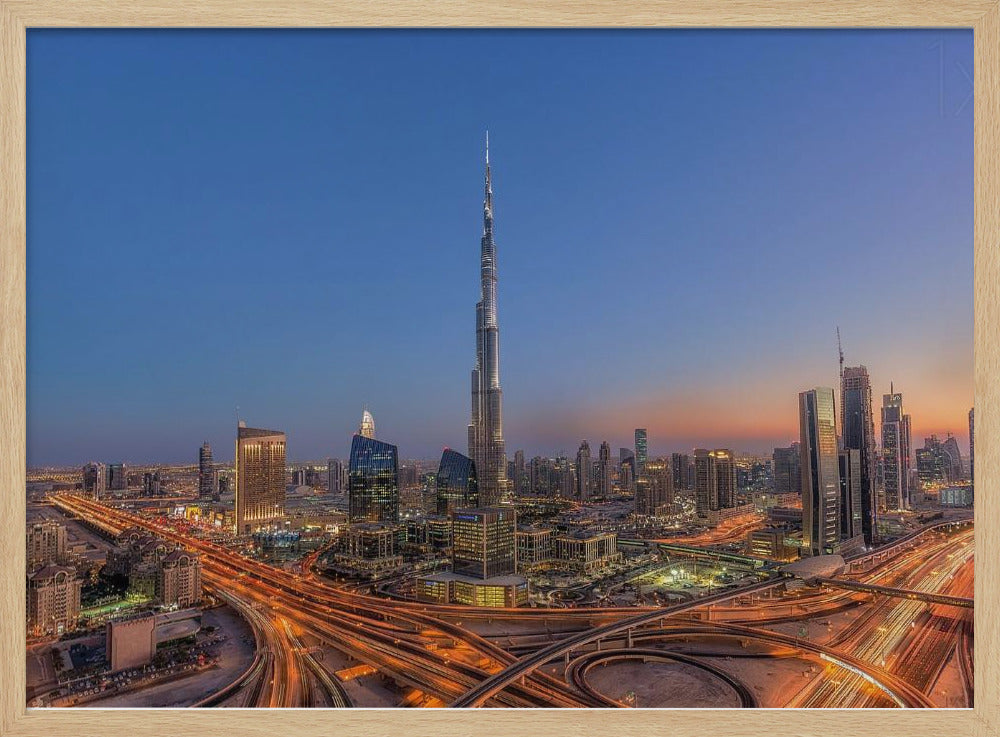An aerial photograph of the Dubai skyline at dusk, centered on the illuminated Burj Khalifa. The sky transitions from deep blue to warm orange at the horizon, while a complex network of highways below glows with the light trails of traffic. Artwork