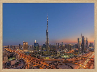An aerial photograph of the Dubai skyline at dusk, centered on the illuminated Burj Khalifa. The sky transitions from deep blue to warm orange at the horizon, while a complex network of highways below glows with the light trails of traffic. Artwork