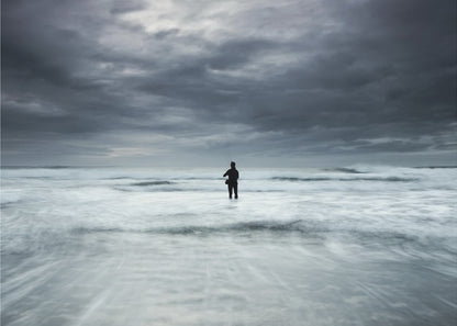 A moody, long-exposure photograph of a silhouetted fisherman standing in the churning white surf of the ocean under a dark, stormy sky, presented in a silver frame. Wall Art