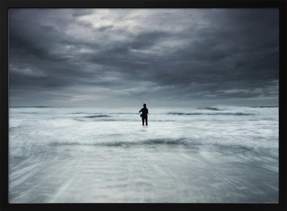 A moody, long-exposure photograph of a silhouetted fisherman standing in the churning white surf of the ocean under a dark, stormy sky, presented in a silver frame. Wall Art