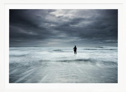 A moody, long-exposure photograph of a silhouetted fisherman standing in the churning white surf of the ocean under a dark, stormy sky, presented in a silver frame. Wall Art