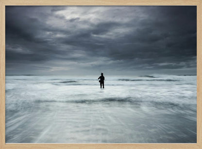 A moody, long-exposure photograph of a silhouetted fisherman standing in the churning white surf of the ocean under a dark, stormy sky, presented in a silver frame. Wall Art