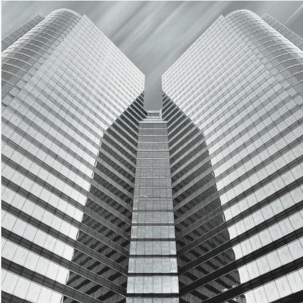 A low-angle, symmetrical black and white photograph of a modern glass skyscraper. The two towers of the building converge towards the center, creating a V-shape against a sky with motion-blurred clouds. The photo is enclosed in a light-colored wooden frame. Print