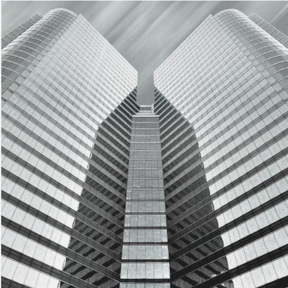 A low-angle, symmetrical black and white photograph of a modern glass skyscraper. The two towers of the building converge towards the center, creating a V-shape against a sky with motion-blurred clouds. The photo is enclosed in a light-colored wooden frame. Print