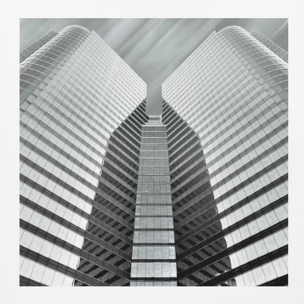 A low-angle, symmetrical black and white photograph of a modern glass skyscraper. The two towers of the building converge towards the center, creating a V-shape against a sky with motion-blurred clouds. The photo is enclosed in a light-colored wooden frame. Print