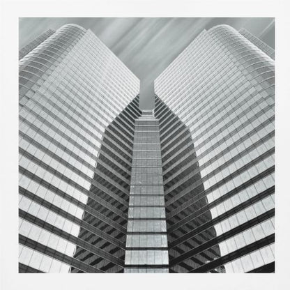 A low-angle, symmetrical black and white photograph of a modern glass skyscraper. The two towers of the building converge towards the center, creating a V-shape against a sky with motion-blurred clouds. The photo is enclosed in a light-colored wooden frame. Print