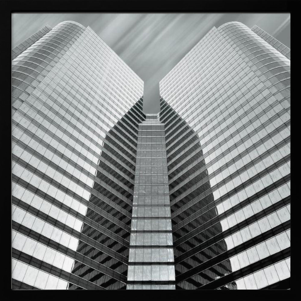 A low-angle, symmetrical black and white photograph of a modern glass skyscraper. The two towers of the building converge towards the center, creating a V-shape against a sky with motion-blurred clouds. The photo is enclosed in a light-colored wooden frame. Print