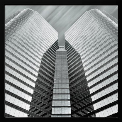 A low-angle, symmetrical black and white photograph of a modern glass skyscraper. The two towers of the building converge towards the center, creating a V-shape against a sky with motion-blurred clouds. The photo is enclosed in a light-colored wooden frame. Print