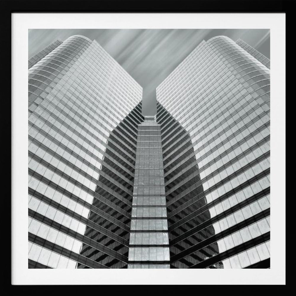 A low-angle, symmetrical black and white photograph of a modern glass skyscraper. The two towers of the building converge towards the center, creating a V-shape against a sky with motion-blurred clouds. The photo is enclosed in a light-colored wooden frame. Print