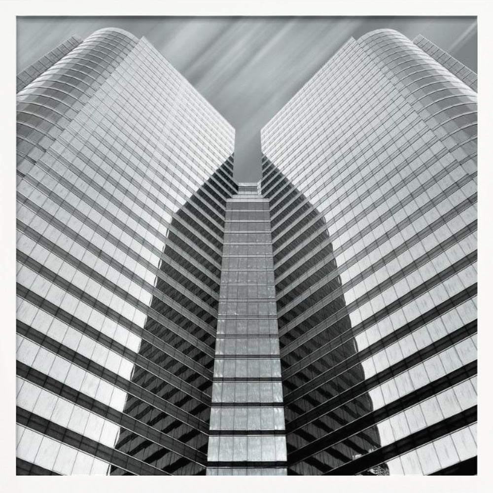 A low-angle, symmetrical black and white photograph of a modern glass skyscraper. The two towers of the building converge towards the center, creating a V-shape against a sky with motion-blurred clouds. The photo is enclosed in a light-colored wooden frame. Print