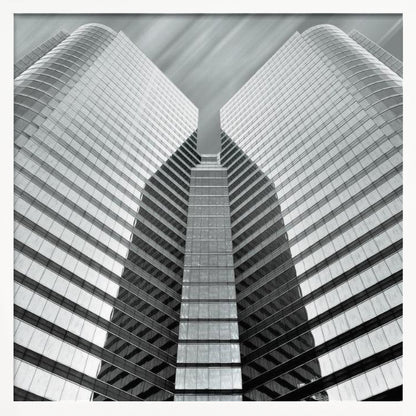 A low-angle, symmetrical black and white photograph of a modern glass skyscraper. The two towers of the building converge towards the center, creating a V-shape against a sky with motion-blurred clouds. The photo is enclosed in a light-colored wooden frame. Print