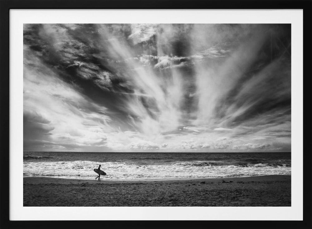 A dramatic black and white photograph of a lone surfer silhouetted against the sea, walking along a sandy beach. The sky is filled with spectacular, radiating clouds that stretch across the horizon, creating a powerful and moody atmosphere. The image is enclosed in a silver frame. Poster