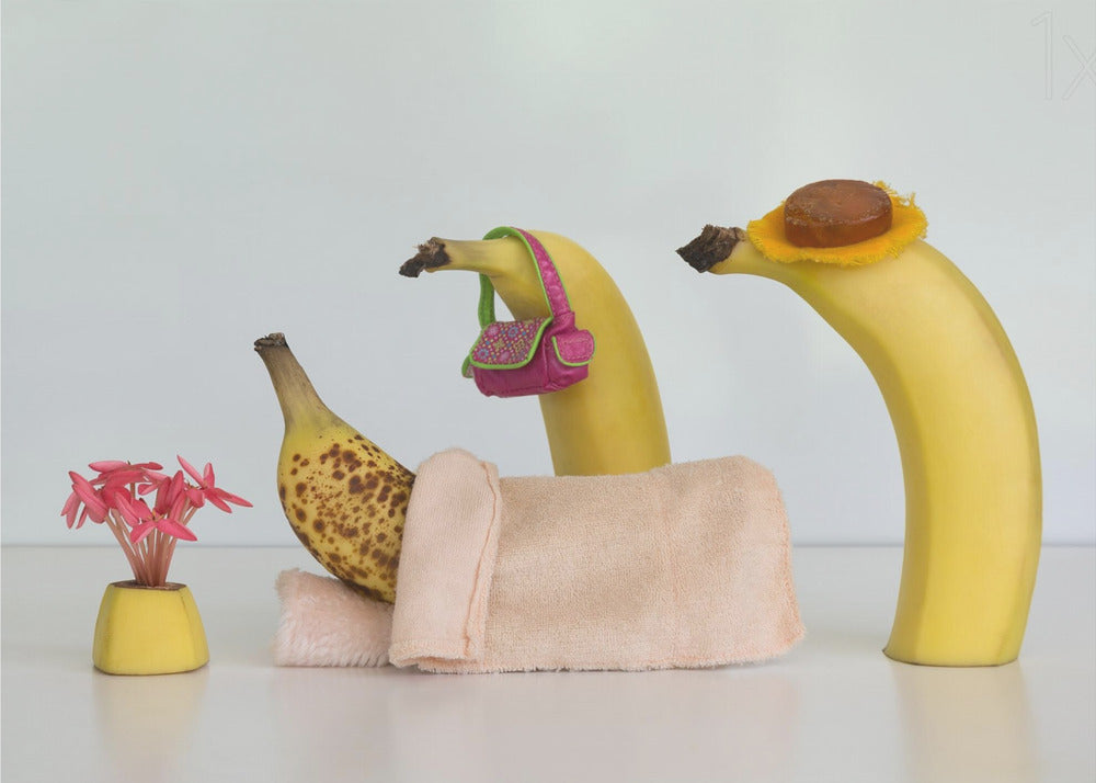 A whimsical still life photo featuring three bananas personified as a family or friends. One overripe, spotted banana lies down, covered by a pink towel, while another banana with a tiny pink purse and a third with a small hat seem to be visiting. A small vase with pink flowers sits nearby on the white surface. Poster