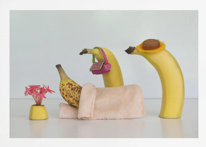 A whimsical still life photo featuring three bananas personified as a family or friends. One overripe, spotted banana lies down, covered by a pink towel, while another banana with a tiny pink purse and a third with a small hat seem to be visiting. A small vase with pink flowers sits nearby on the white surface. Poster