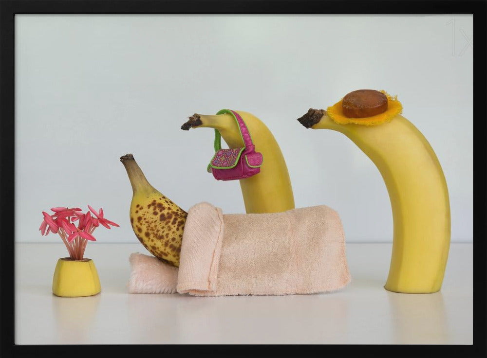 A whimsical still life photo featuring three bananas personified as a family or friends. One overripe, spotted banana lies down, covered by a pink towel, while another banana with a tiny pink purse and a third with a small hat seem to be visiting. A small vase with pink flowers sits nearby on the white surface. Poster