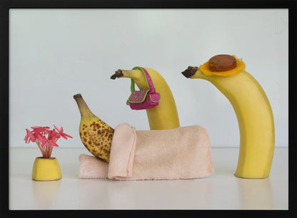 A whimsical still life photo featuring three bananas personified as a family or friends. One overripe, spotted banana lies down, covered by a pink towel, while another banana with a tiny pink purse and a third with a small hat seem to be visiting. A small vase with pink flowers sits nearby on the white surface. Poster