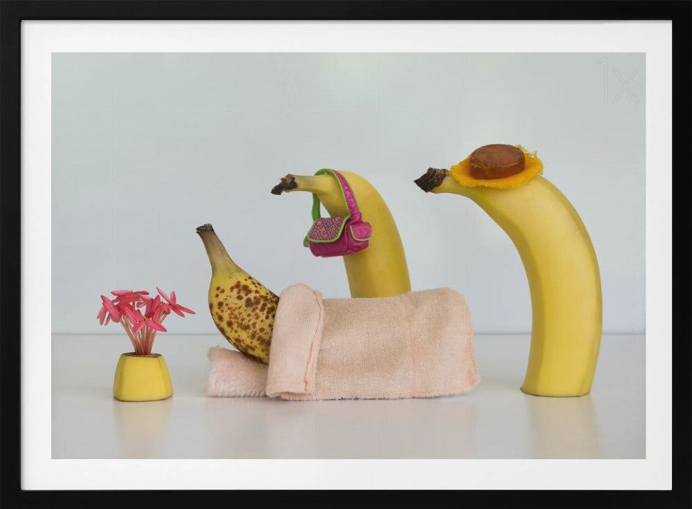 A whimsical still life photo featuring three bananas personified as a family or friends. One overripe, spotted banana lies down, covered by a pink towel, while another banana with a tiny pink purse and a third with a small hat seem to be visiting. A small vase with pink flowers sits nearby on the white surface. Poster