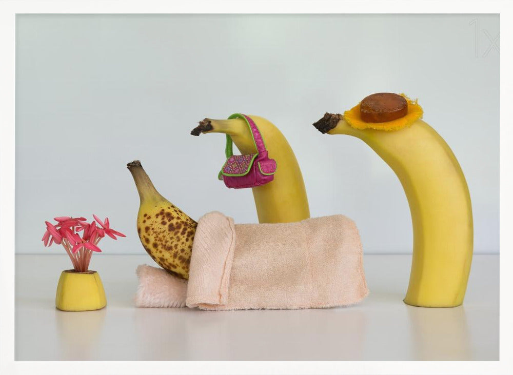 A whimsical still life photo featuring three bananas personified as a family or friends. One overripe, spotted banana lies down, covered by a pink towel, while another banana with a tiny pink purse and a third with a small hat seem to be visiting. A small vase with pink flowers sits nearby on the white surface. Poster