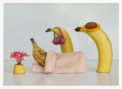 A whimsical still life photo featuring three bananas personified as a family or friends. One overripe, spotted banana lies down, covered by a pink towel, while another banana with a tiny pink purse and a third with a small hat seem to be visiting. A small vase with pink flowers sits nearby on the white surface. Poster