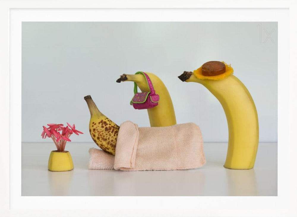 A whimsical still life photo featuring three bananas personified as a family or friends. One overripe, spotted banana lies down, covered by a pink towel, while another banana with a tiny pink purse and a third with a small hat seem to be visiting. A small vase with pink flowers sits nearby on the white surface. Poster