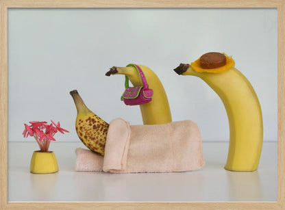 A whimsical still life photo featuring three bananas personified as a family or friends. One overripe, spotted banana lies down, covered by a pink towel, while another banana with a tiny pink purse and a third with a small hat seem to be visiting. A small vase with pink flowers sits nearby on the white surface. Poster