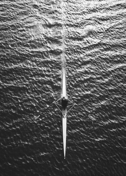 A dramatic black and white overhead photograph of a lone rower in a sleek racing shell, cutting a straight path through dark, textured water. The high-contrast lighting highlights the ripples on the water's surface and the rower's focused form. Artwork