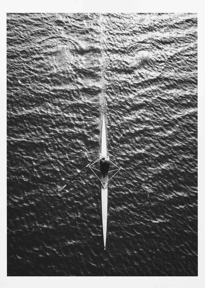 A dramatic black and white overhead photograph of a lone rower in a sleek racing shell, cutting a straight path through dark, textured water. The high-contrast lighting highlights the ripples on the water's surface and the rower's focused form. Artwork
