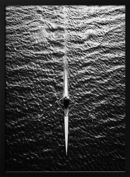 A dramatic black and white overhead photograph of a lone rower in a sleek racing shell, cutting a straight path through dark, textured water. The high-contrast lighting highlights the ripples on the water's surface and the rower's focused form. Artwork