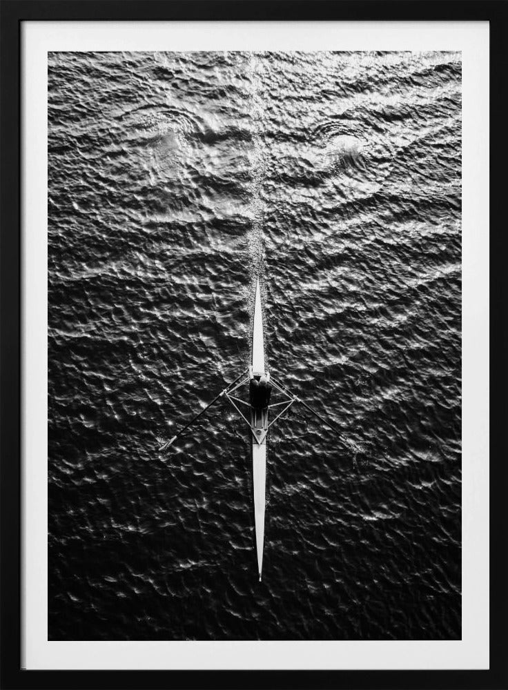 A dramatic black and white overhead photograph of a lone rower in a sleek racing shell, cutting a straight path through dark, textured water. The high-contrast lighting highlights the ripples on the water's surface and the rower's focused form. Artwork